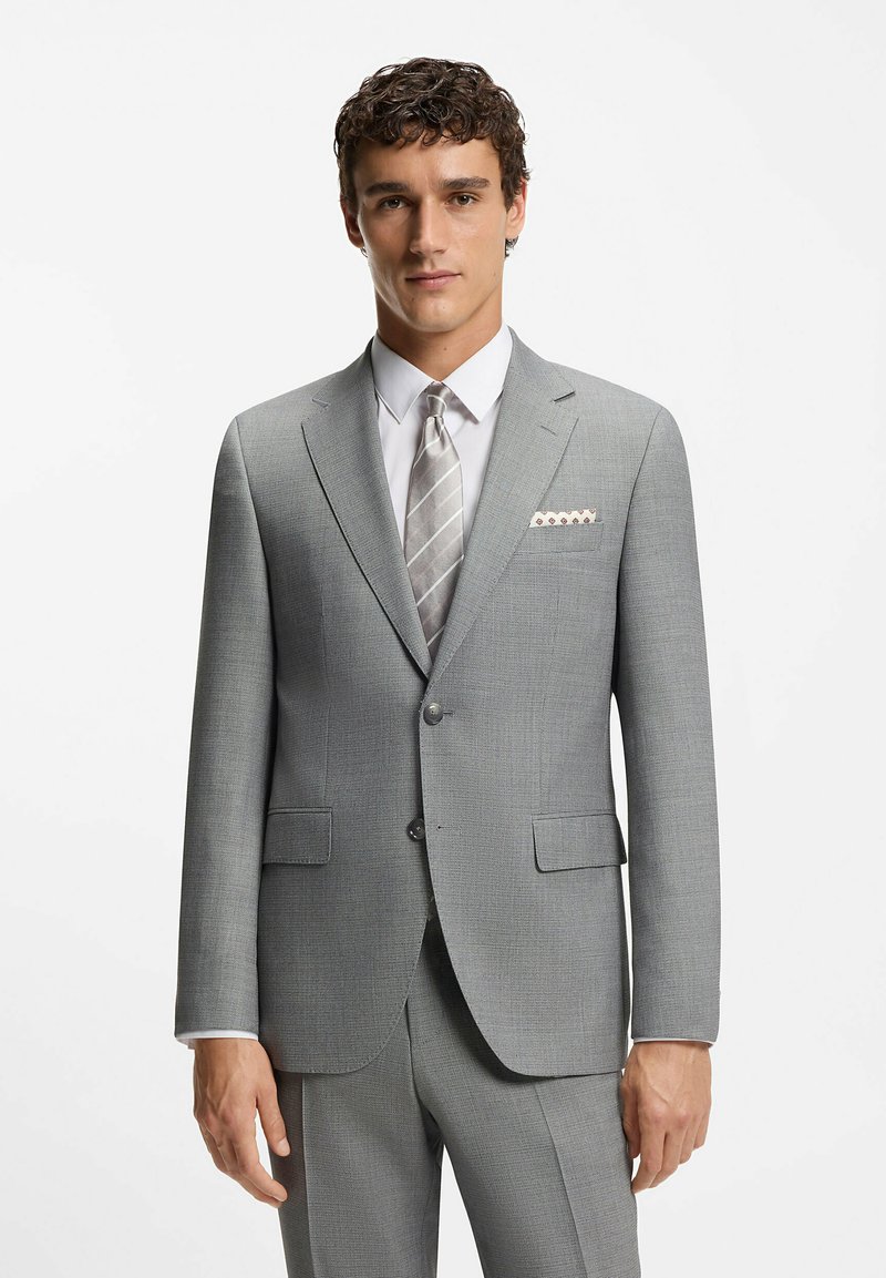 Uomo che indossa un completo grigio chiaro con una camicia bianca, una cravatta a righe argentate e un fazzoletto da taschino a fantasia, in piedi contro uno sfondo bianco semplice.