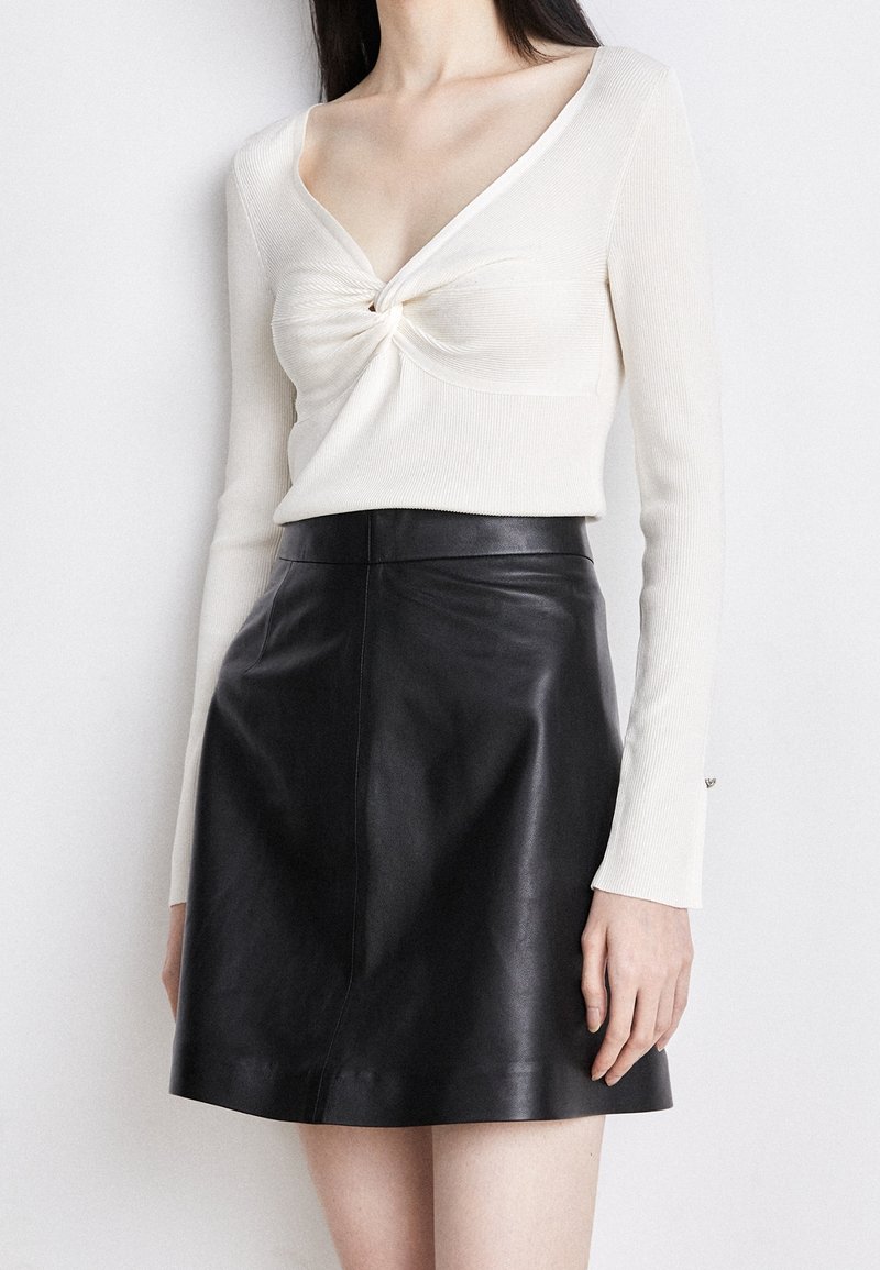 Top a maniche lunghe a coste crema con un davanti annodato; abbinato a una mini gonna in pelle nera, texture liscia, design aderente.