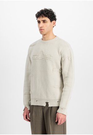 Mand med kort krøllet hår iført en lys beige slidt striktrøje og løse grå bukser mod en ensfarvet hvid baggrund.