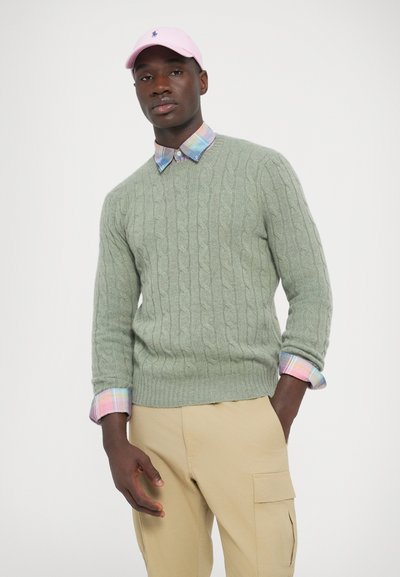 Camisola verde-claro de malha com mangas até meio braço, usada sobre uma camisa com gola aos quadrados; combinada com calças cargo bege e um boné rosa.