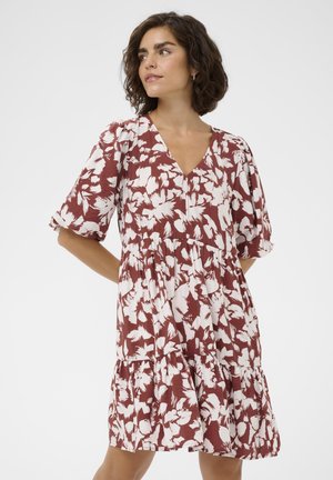 Kvinde med kort krøllet hår iført en løs brun kjole med hvide blomsterprint, stående med hænderne bag ryggen.