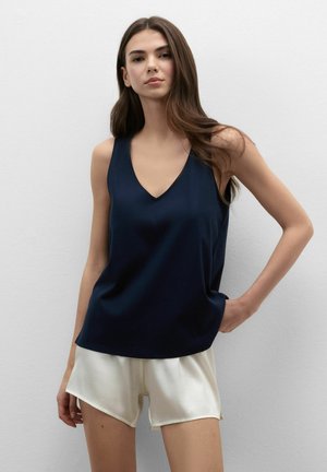 Frau mit langen braunen Haaren, die ein ärmelloses marineblaues Top und weiße Shorts trägt, steht vor einer schlichten hellen Wand.