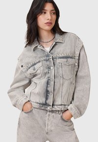 Giacca in denim grigia con hardware argentato, vestibilità oversize e sbiaditure distintive. Presenta grandi tasche anteriori e rivetti decorativi lungo le spalle.