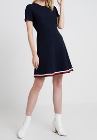 Vestido corto de color navy con mangas cortas y falda acampanada, con un dobladillo a rayas en blanco y rojo. Combinado con botines blancos hasta el tobillo. Tejido suave, con corsé ajustado.