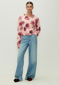 Cardigan rosa con motivo floreale rosso, scollatura rotonda e bottoni. Abbinato a jeans azzurri a gamba larga e pantofole bordeaux.