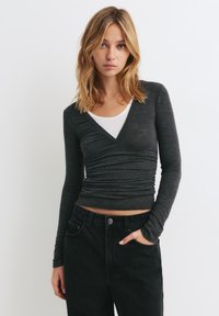 Grå langærmet tætsiddende top med V-hals og rynket tekstur, lagdelt over en hvid top, parret med højtaljede sorte jeans.