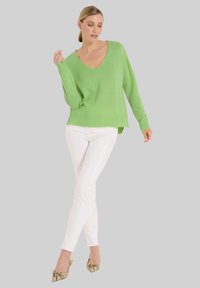 Grüner Strickpullover mit V-Ausschnitt, langen Ärmeln und relaxtem Schnitt, kombiniert mit weißen, engen Hosen und gemusterten spitz zulaufenden Schuhen.