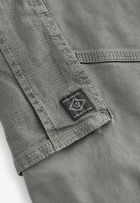 Tejido gris con una textura suave, que presenta un dobladillo cosido y una etiqueta tejida negra con texto blanco y un logotipo en el lado izquierdo.