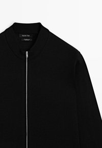 Sort bomberjakke lavet af 100% merino uld. Har høj krave, frontlynlås og ribbede manchetter. Glat tekstur og minimalistisk design.