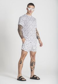 Top blanco de manga corta y pantalones cortos con un patrón repetido de logotipos negros, combinados con sandalias negras y gafas de sol reflectantes.
