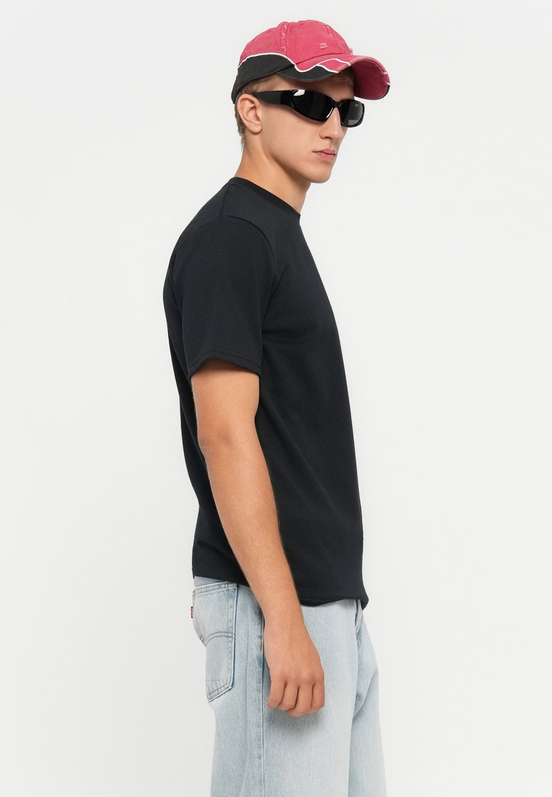 Młody mężczyzna w czarnej koszulce, jasnoniebieskich dżinsach, czerwono-czarnym czapce i czarnych okularach przeciwsłonecznych, stojący bokiem na białym tle.