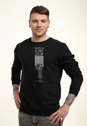 Man met tatoeages in een zwart sweatshirt met een grafisch ontwerp en de tekst "Zwart is mijn gelukkige kleur," staande met zijn handen op zijn heupen tegen een witte achtergrond.