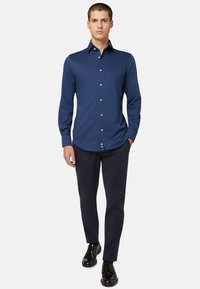 Mornarsko modra srajca z dolgimi rokavi in gumbom na ovratniku, gladka tekstura in bele gumbe, v kombinaciji s temnimi hlačami in črnimi čevlji.