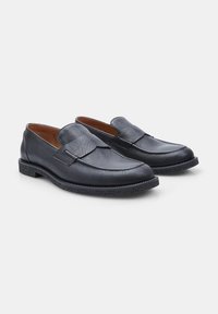 Mocasines de cuero negro con textura suave, punta redondeada y acentos elásticos en los laterales sobre una suela de goma texturizada. Diseño casual adecuado para diversas ocasiones.