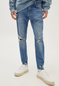 Persona che indossa jeans chiari strappati con buchi sulle ginocchia e sulle cosce, abbinati a scarpe da ginnastica bianche e a un felpa blu.