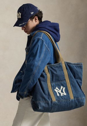 Mladi oseba v modri denim jakni in mornarsko modri kapi, nosi na rami veliko modro denim torbo z belim logotipom NY.