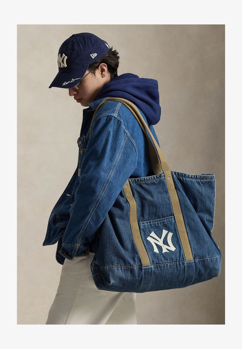 Νεαρός με μπλε τζιν μπουφάν και μπλε καπέλο να κρατά μια μεγάλη μπλε τζιν τσάντα ώμου με λευκό λογότυπο NY.