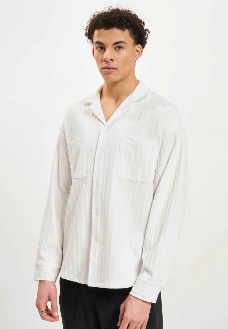 Chemise blanche à boutons, avec des rayures verticales, manches longues, deux poches poitrine et un col décontracté. Tissu doux et lisse avec une texture subtile.