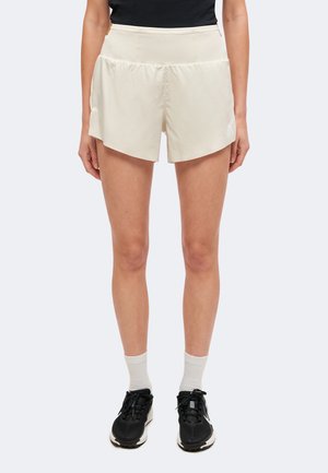 Femme portant un short de sport beige à taille haute, des baskets noires, des chaussettes blanches et un haut noir, debout devant un fond clair uni.