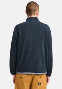Granatowy polarowy sweter z kołnierzem stójkowym i prostokątną metką z tyłu. Wyróżnia się jasnoszarym dolnym obszyciem. Gładka tekstura.