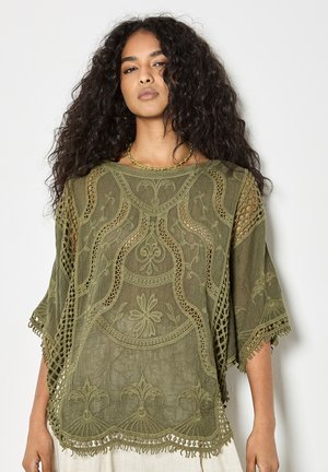 Femme aux longs cheveux bouclés portant un haut en dentelle brodée vert olive à larges manches et un collier de perles, debout devant un fond blanc.