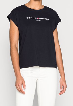 Person, die ein schwarzes ärmelloses Tommy Hilfiger T-Shirt mit Logo und weiße Hose trägt, von Hals bis Hüfte gezeigt, stehend vor einem einfarbigen Hintergrund.