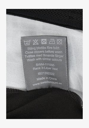 Sort tre-lags vest med en glat grå indvendig foring, med plejeinstruktioner trykt på et stofmærke, sikkert syede kanter.
