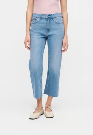 Donna che indossa jeans corti a vita alta azzurri con orli grezzi, abbinati a scarpe basse beige con lacci e top rosa chiaro senza maniche.