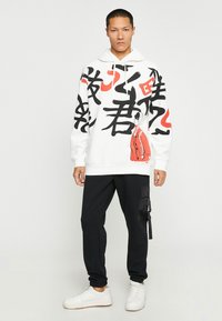 Biała bluza z kapturem z czarnymi i czerwonymi grafikami, z wyrazistym tekstem i postacią. Połączona z czarnymi zwężanymi spodniami i białymi sneakersami.