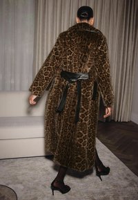 Pitkä, ruskea leopardikuosisesta tekoturkiksesta valmistettu takki, jossa on musta vyöllinen vyötärö, leveä kaulus ja löysät hihat. Taustalla pehmeä sisustus.