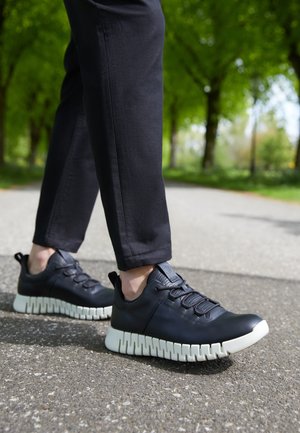 Schwarze Ledersneaker mit strukturierter Oberfläche, ausgestattet mit einer klobigen weißen Sohle und elastischen Schnürsenkeln, vor einem grauen Weg und grünen Bäumen.