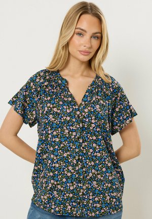 Mujer rubia con blusa de manga corta abotonada con un patrón floral azul, rosa y verde, de pie con las manos detrás de la espalda.