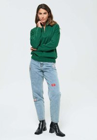 Un sweat zippé vert associé à un jean bleu clair avec des patchs. Des bottes à talons noires complètent la tenue. Fond simple.