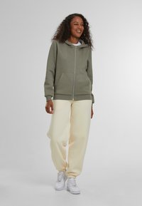 Sudadera con cremallera de color verde claro, con bolsillos delanteros, combinada con pantalones de chándal sueltos de color amarillo pálido y zapatillas blancas. Tejido texturizado, diseño informal.