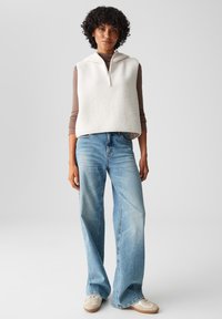 Weißer Fleece-Westen mit Kapuze und Reißverschluss, über einem braunen Langarmshirt getragen. Hellblaue Weite-Bein-Jeans und beigefarbene Sneakers runden das Outfit ab.