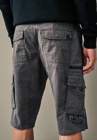 Graue Cargo-Shorts aus leichtem Material, ausgestattet mit mehreren Taschen, einer seitlichen Reißverschlusstasche und gesteppten Details auf der Rückseite.