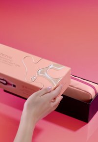 Hand hält eine rosa Schachtel mit transparentem Design und setzt sie auf eine schwarze offene Schachtel, die eine rosa Reißverschlusstasche auf einer rosa Oberfläche enthält.