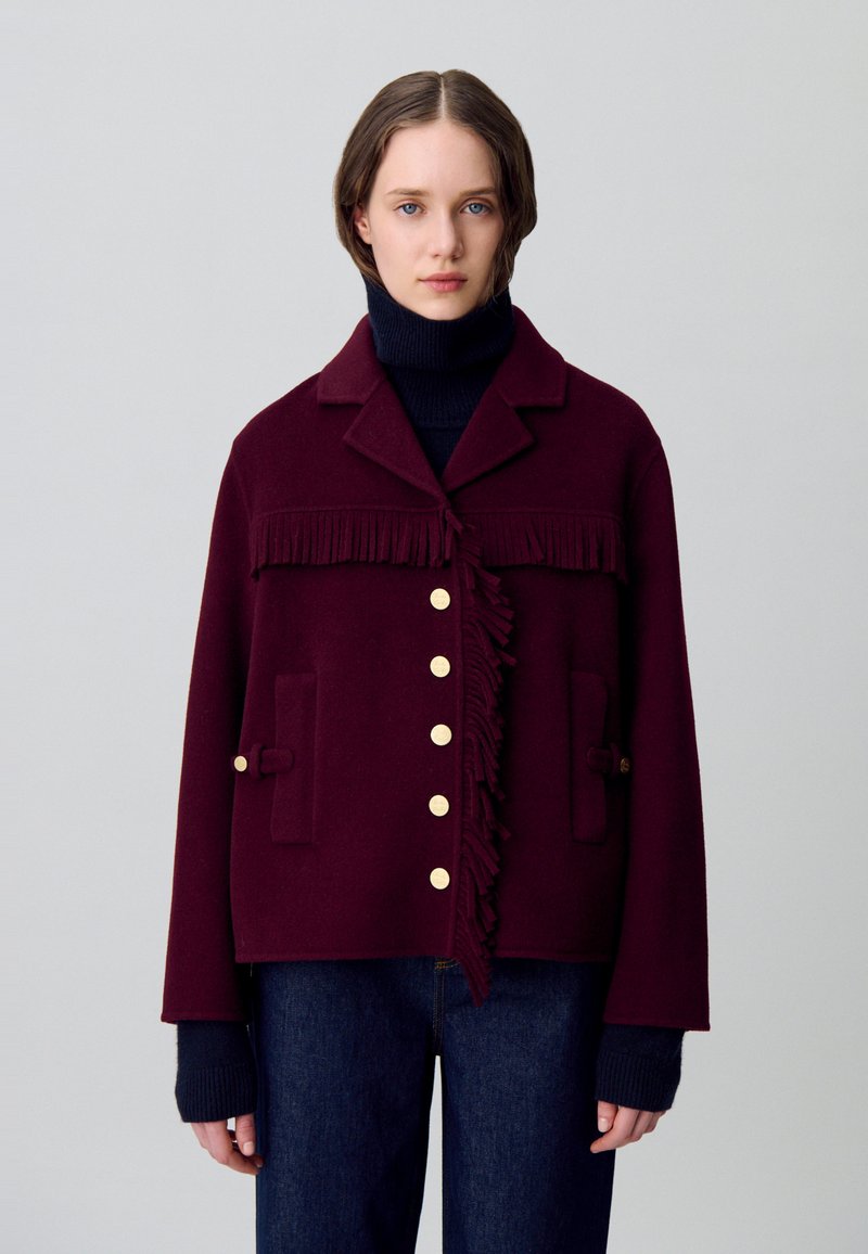 Chaqueta de lana burdeos con detalle de flecos, botones dorados y bolsillos de parche. Combinada con un suéter de cuello alto oscuro y jeans azules.