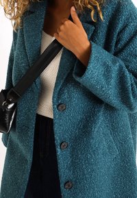 Cappotto peloso turchese con finitura strutturata, caratterizzato da grandi bottoni neri e colletto con risvolto. Abbinato a una maglia bianca lavorata a maglia e jeans scuri.