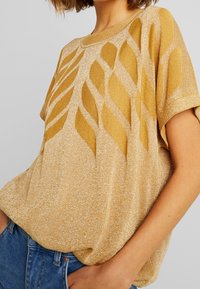 Mujer vestida con una blusa de punto dorada con brillo, de mangas cortas y motivos de hojas, combinada con jeans de mezclilla azules, con la mano en el bolsillo.