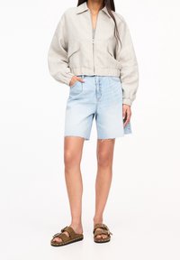 Veste bomber en lin gris clair avec une fermeture zippée devant, associée à un short en denim bleu clair taille haute et des sandales mule marron cloutées.
