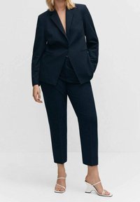 Blazer bleu marine cintré avec revers à cran et un seul bouton. Pantalon assorti raccourci. Sandales blanches à talons avec un talon bloc incurvé.