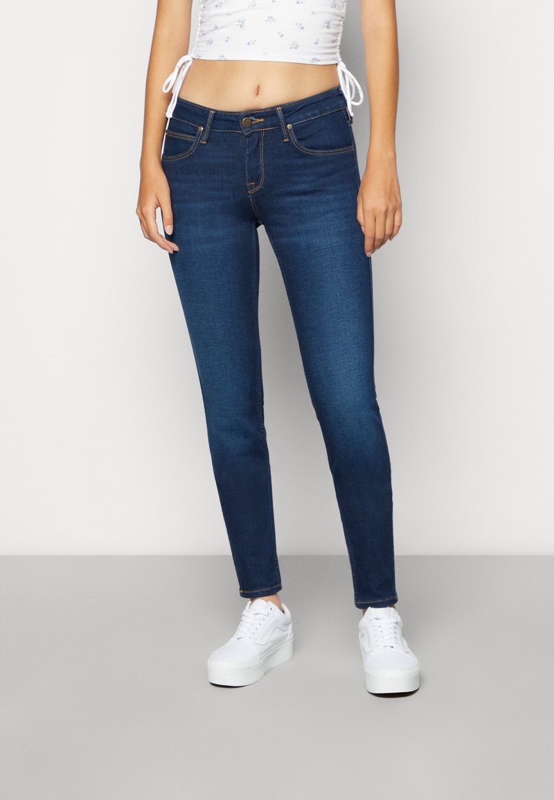 Vaqueros ajustados de mezclilla azul oscuro con cintura de tiro medio, diseño de cinco bolsillos y costuras en contraste, combinados con zapatillas blancas y un top corto.