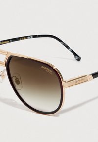 Gafas de sol Carrera con marco dorado, lentes marrones degradados y puntas de patilla negras con patrón, descansando sobre una superficie blanca.