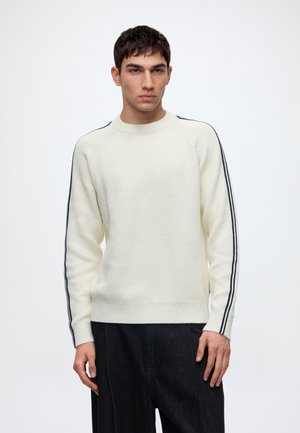 Jeune homme portant un pull en maille crème avec des rayures noires sur les manches et un pantalon noir ample sur fond blanc.