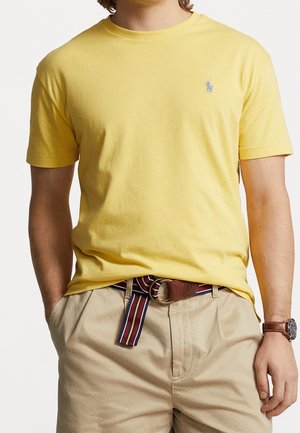 Man draagt een geel shirt met korte mouwen en een klein logo, beige broek, een gestreepte stoffen riem en een polshorloge aan de linkerarm.