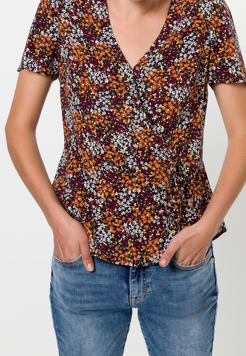 Top floral con mangas cortas, con una base color burdeos oscuro y flores multicolores en naranja, blanco y verde. Diseño peplum.