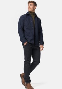 Marinblå struktureret jakke med knaplukning, sidelommer, båret over en grøn turtleneck, sammen med sorte bukser og brune ankelstøvler.