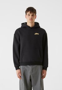 Črna kapuca iz mehkega blaga, ki ima sprednji kenguru žep in zlat logotip na levi prsni strani. Ohlapen kroj z rebrastimi manšeti.