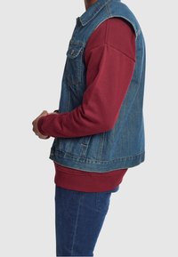 Denimväst med axelsömmar, tillsammans med en långärmad bordeauxfärgad sweatshirt. Blå jeans är synliga, med ribbade muddar och midjeband.